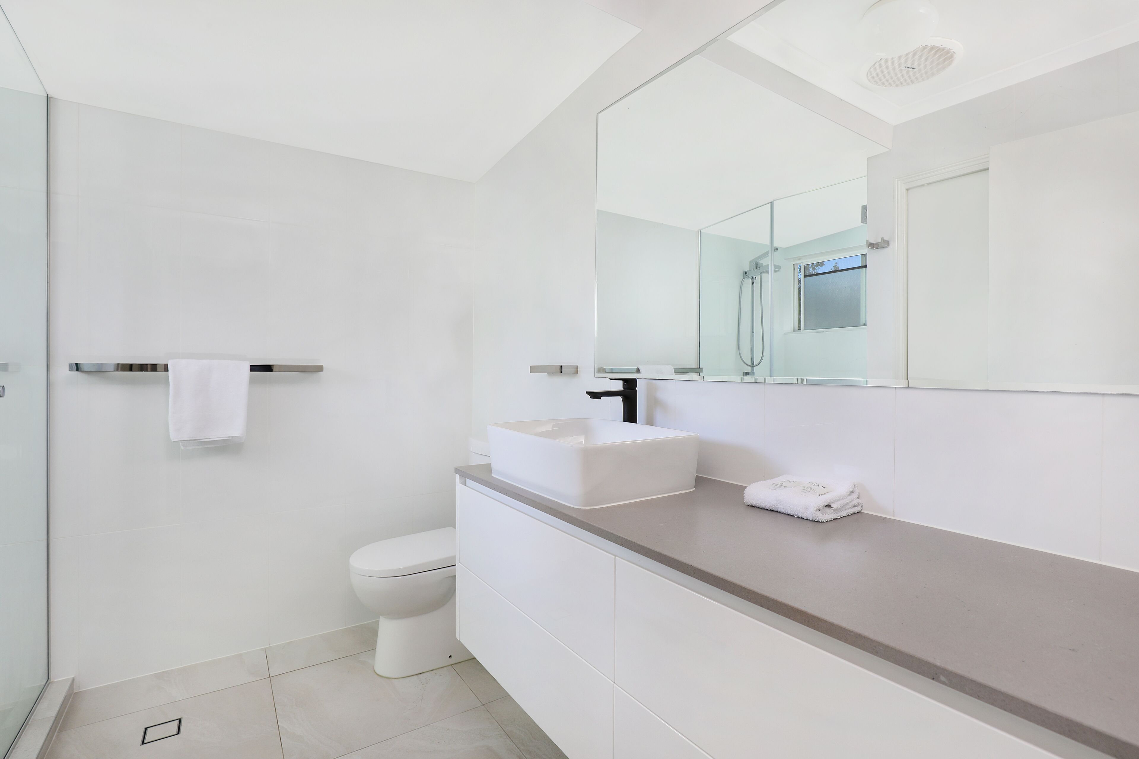 Une salle de bain moderne avec des toilettes blanches, un grand miroir et un comptoir de couleur beige.