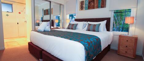 A hotel room with a large bed, a wooden nightstand, a mirror, and a window with a view of trees.