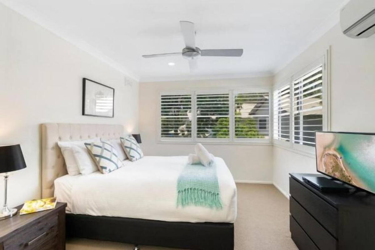 Un dormitorio con cama, ventilador de techo, ventana con persianas, cómoda y televisor.