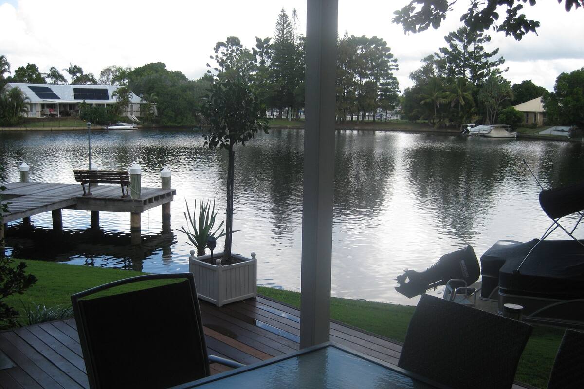 Una terraza con mesa y sillas que da a un lago tranquilo, rodeada de vegetación y algunos edificios al fondo.