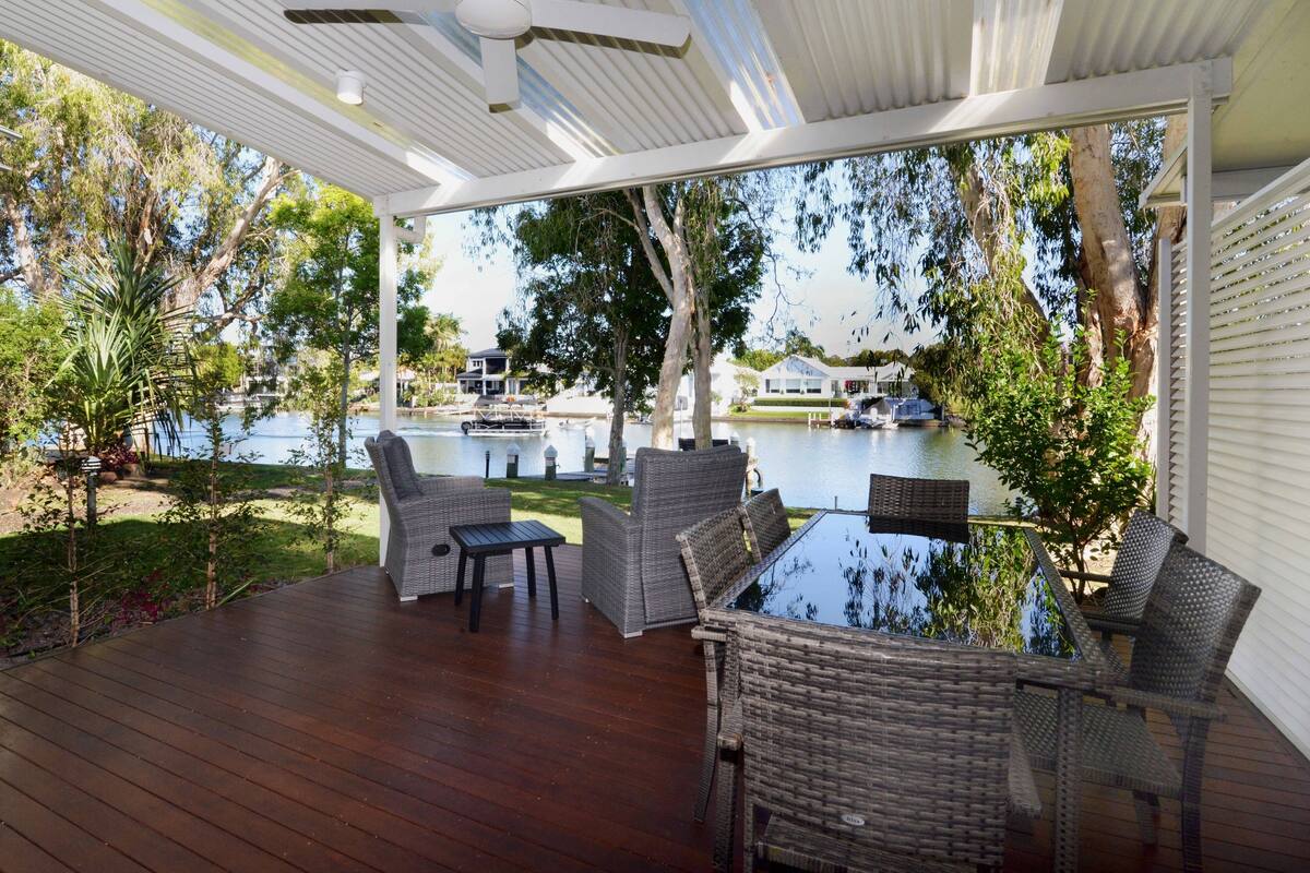 Una terraza cubierta al aire libre con mobiliario de mimbre, una mesa y vistas a un lago y barcos.
