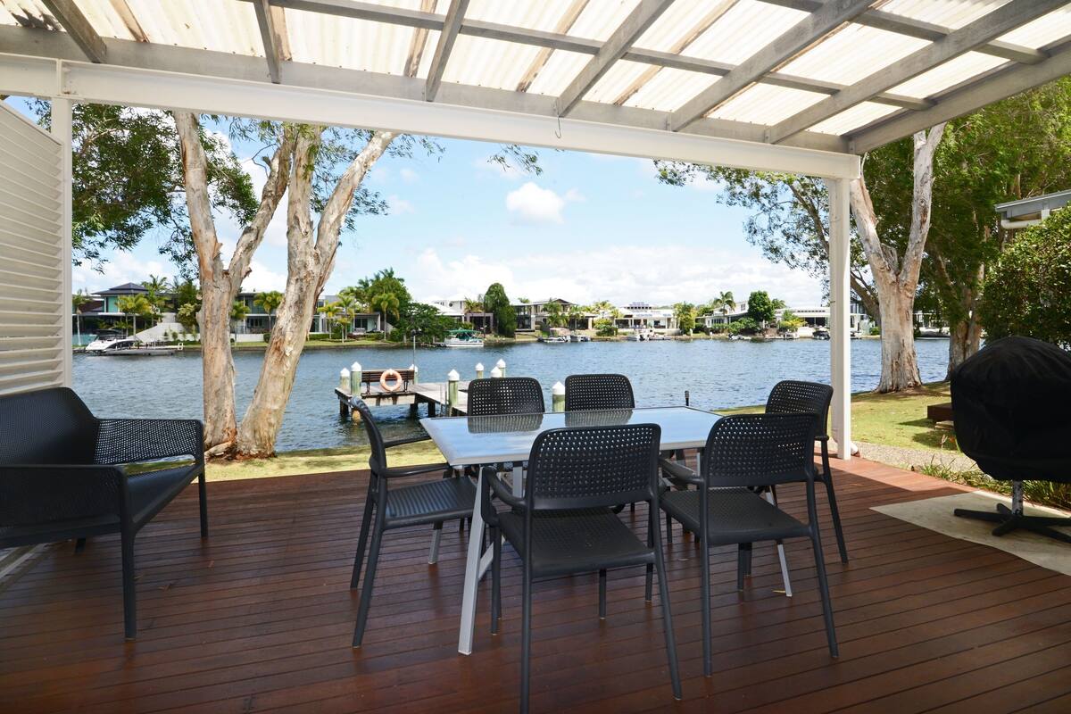 Un espacio de comedor al aire libre cubierto, con vistas a un lago y edificios al fondo.