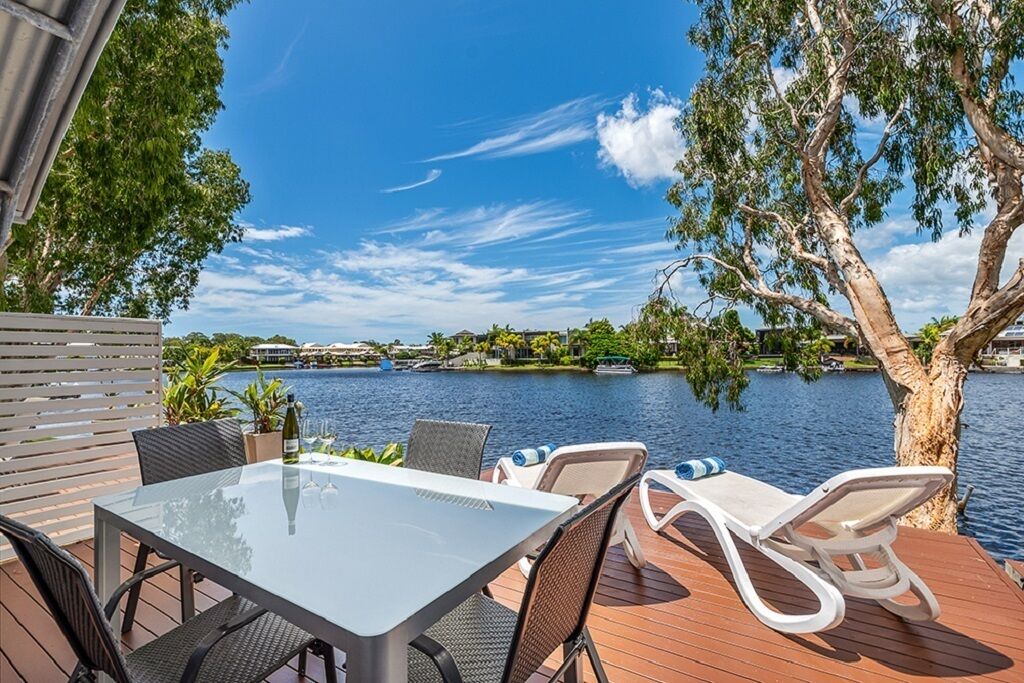 An outdoor deck with a glass table and chairs, overlooking a body of water with trees and buildings in the background.