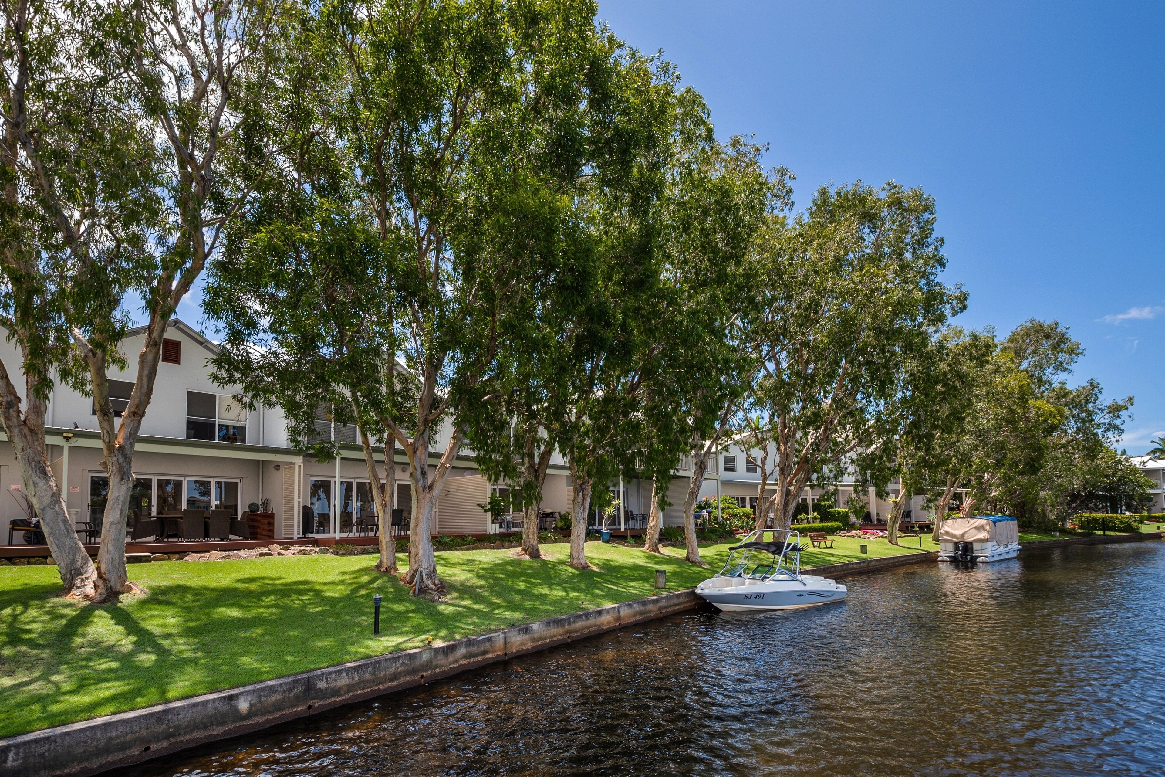 A riverside view with a boat docked on the water, trees lining the path, and a building with a lawn and outdoor seating area.