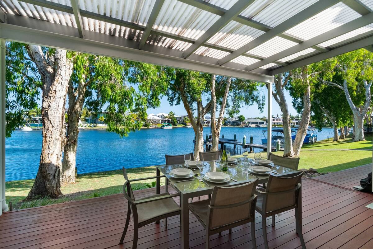 Un espacio para comer al aire libre con vistas a un lago y barcos, rodeado de árboles y un porche cubierto.
