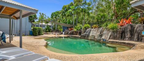 Un área de piscina con sillones de descanso, una zona de estar techada y un paisaje natural de árboles y rocas.