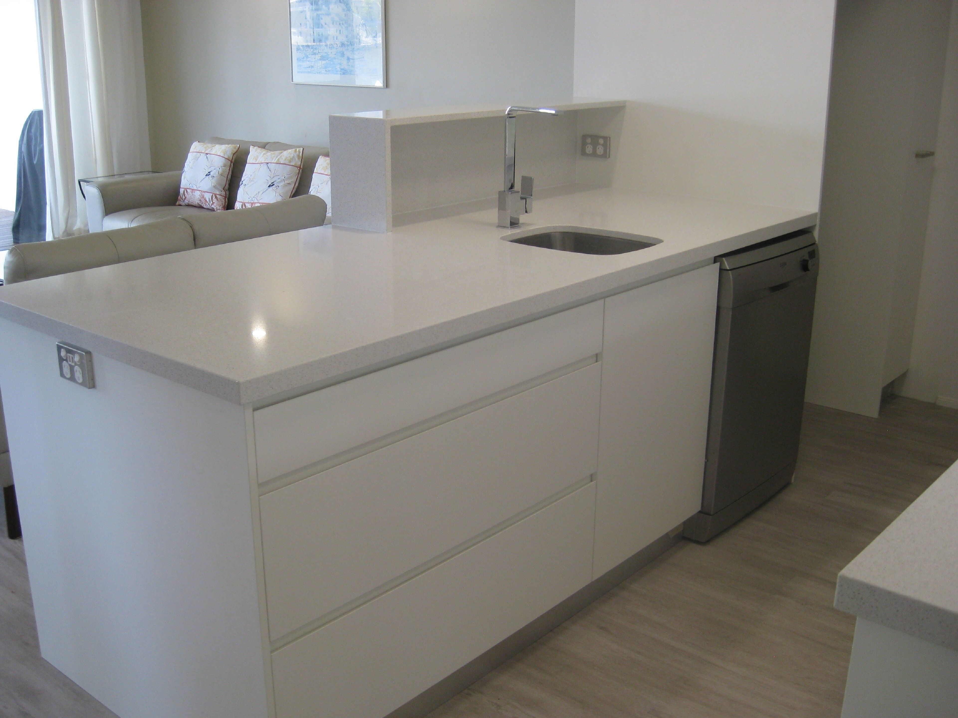 A modern kitchen with a built-in dishwasher, a white countertop, and a grey sofa with patterned cushions.