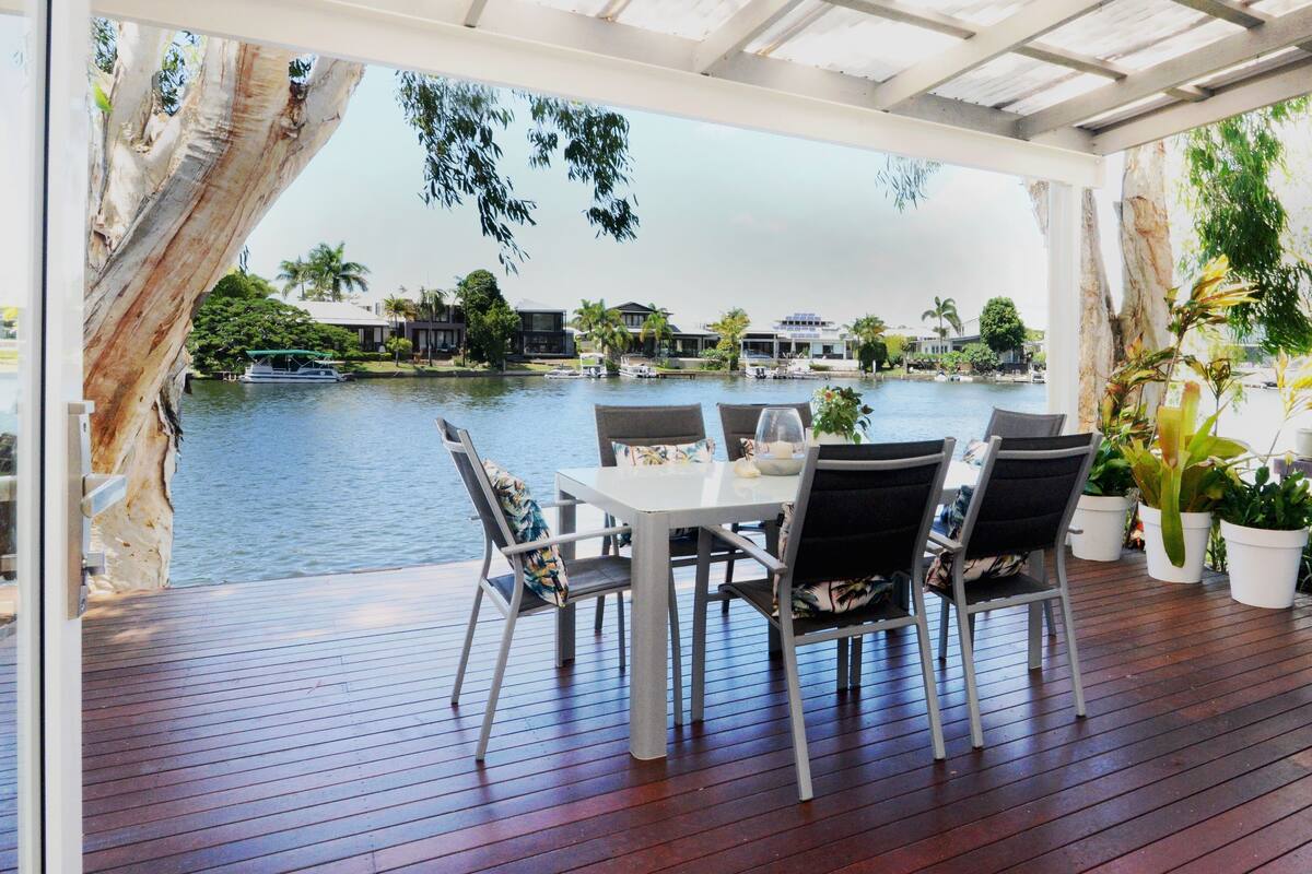Un área de comedor al aire libre cubierta con una mesa y sillas, con vista a un cuerpo de agua con barcos y edificios al fondo.