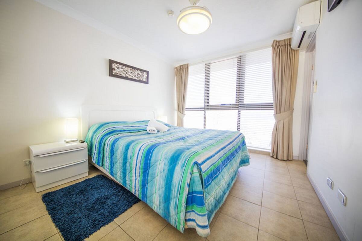 A bedroom with a bed, a nightstand, a lamp, a window with curtains, and an air conditioning unit.