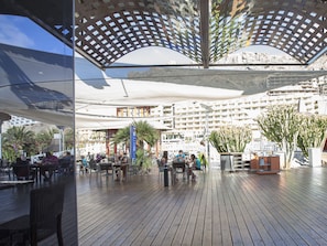 An outdoor deck with wooden flooring, tables, and chairs, surrounded by potted plants and a large umbrella structure.