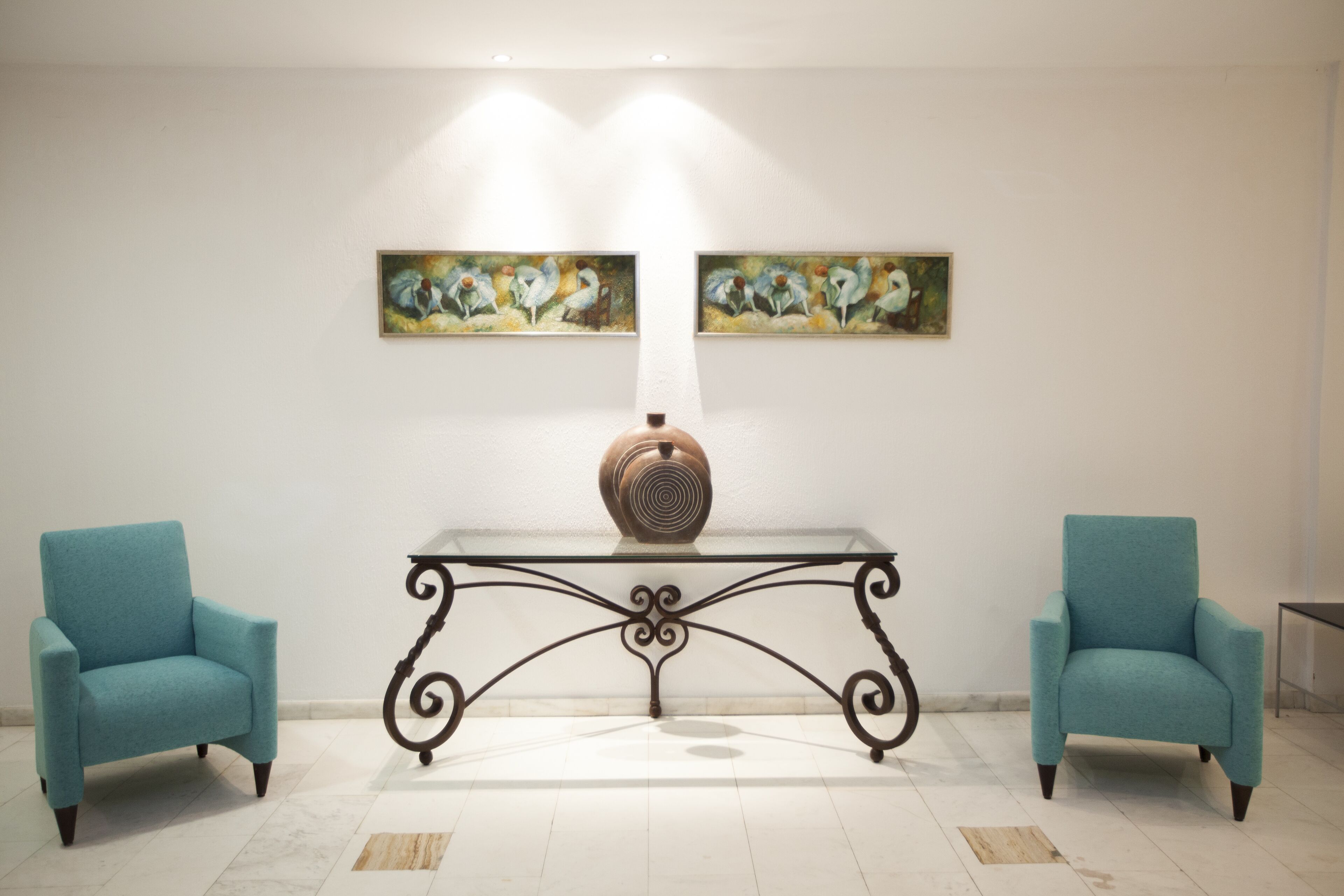 Une pièce comprenant deux fauteuils de couleur turquoise, une table avec un plateau en verre surmontée d’un vase décoratif, et deux tableaux accrochés au mur.