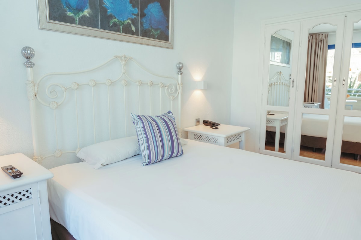 A neatly arranged bedroom with a white bed, a striped pillow, a bedside table with a remote control, and a mirror reflecting a balcony with curtains.