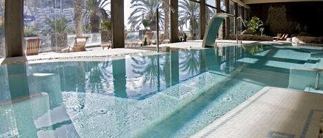 An indoor swimming pool with clear water, large windows, and a view of palm trees and a rocky landscape.