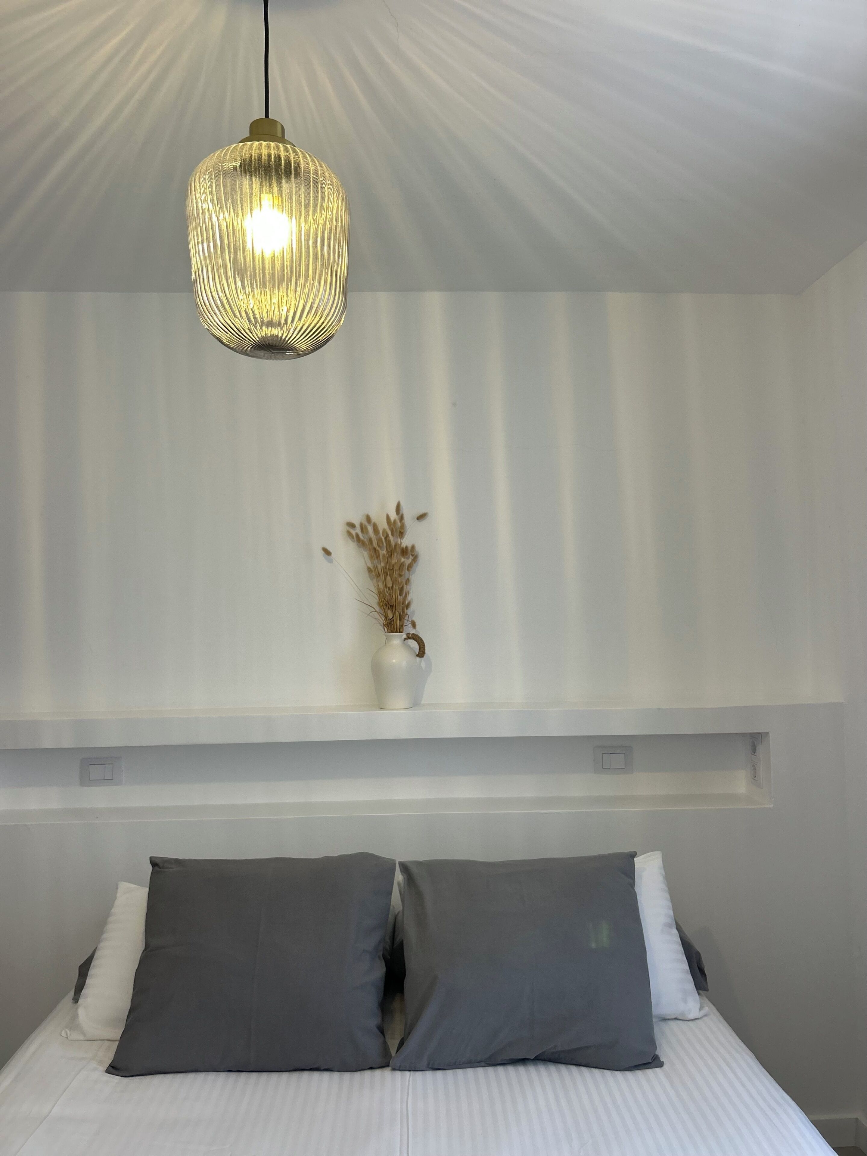 Un dormitorio con cama, almohadas, una lámpara colgante y un florero con plantas secas sobre una repisa.