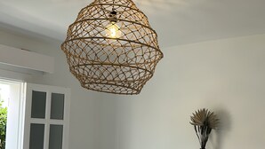 Ein Schlafzimmer mit Bett, Kissen, einem Nachttisch, einer Hängeleuchte und einem Fenster mit Jalousien.