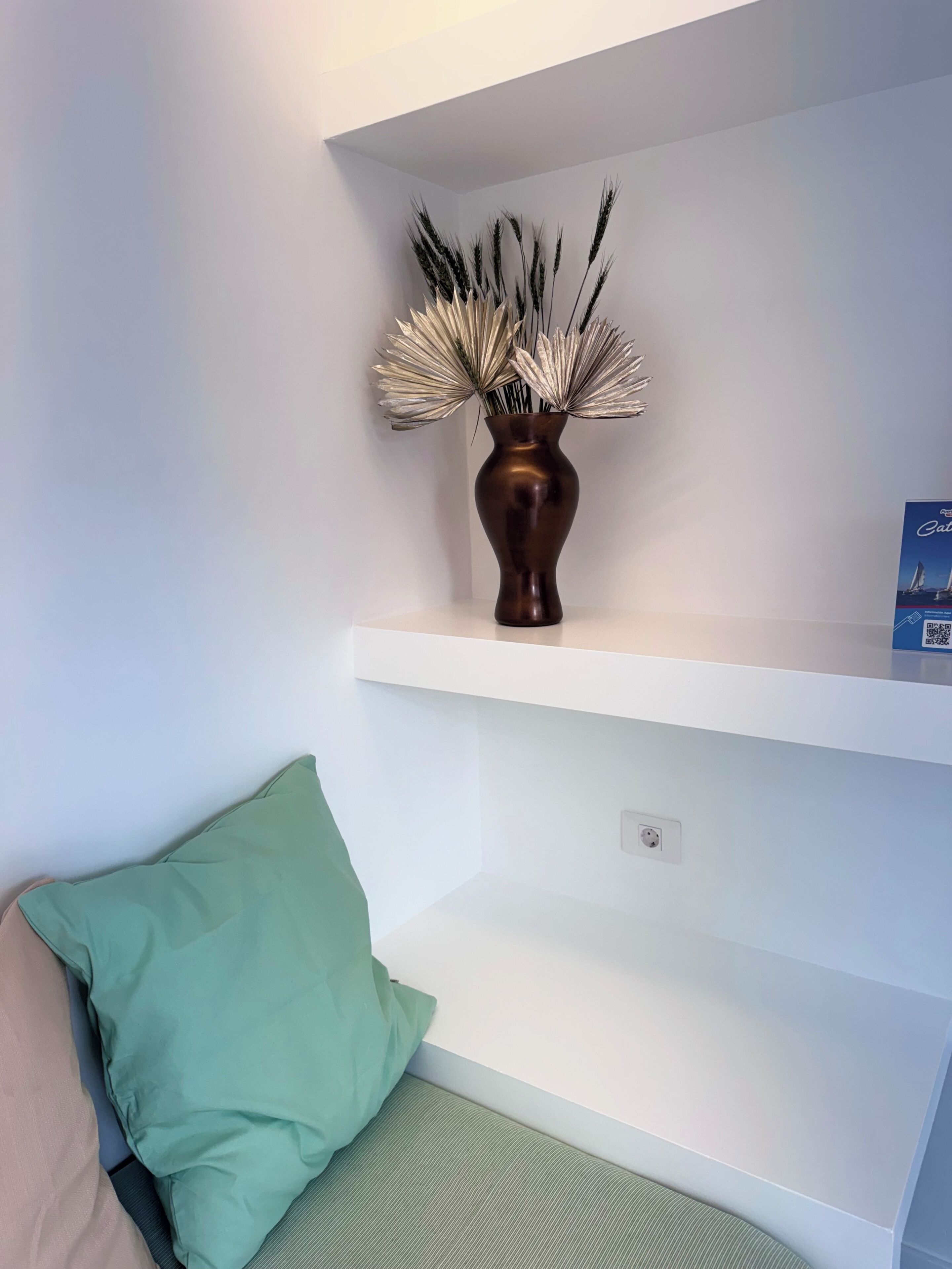 A white wall with a brown vase containing dried plants on a white shelf.