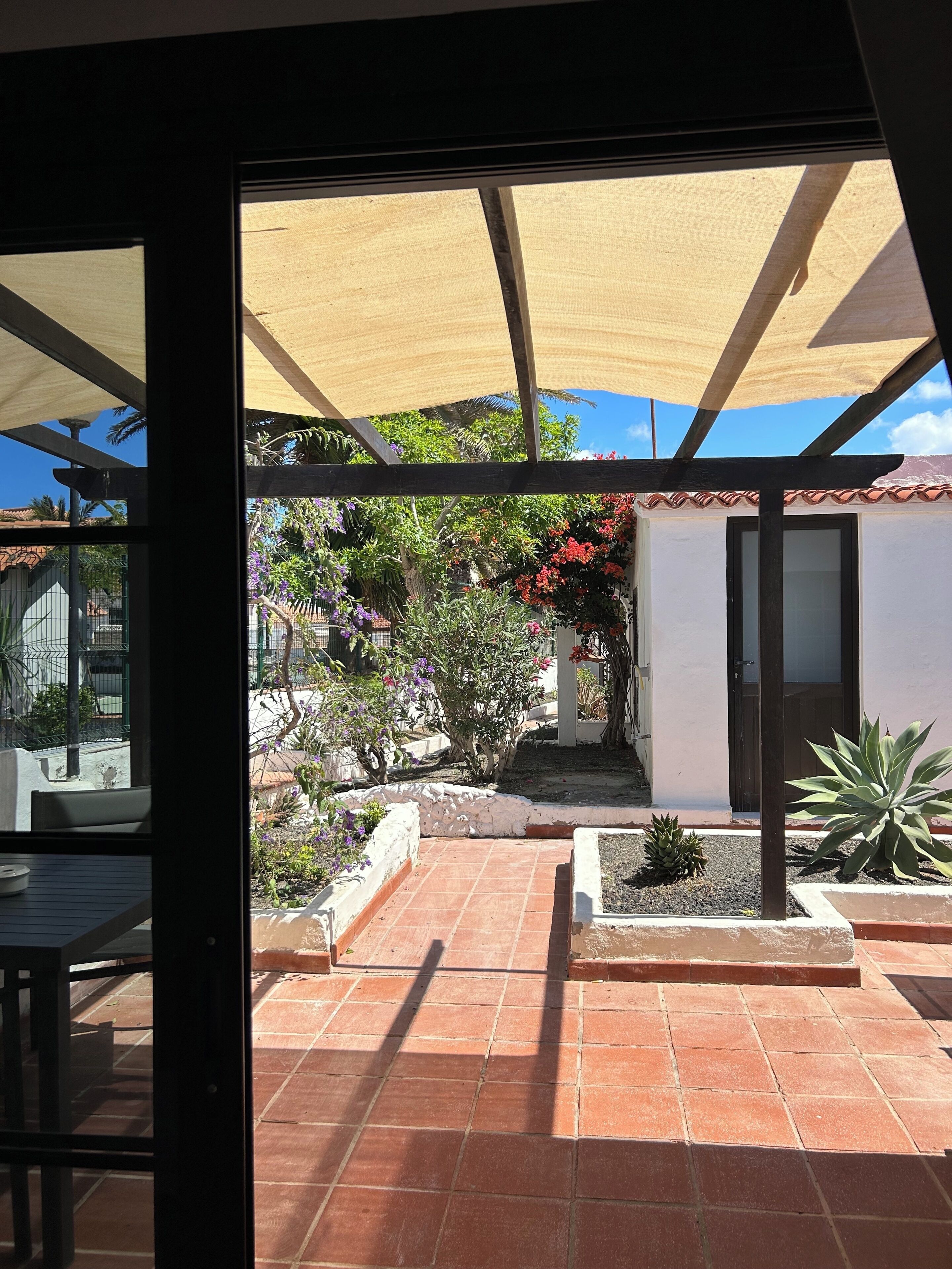 A patio with a tiled floor, a pergola with a shade sail, and various potted plants.