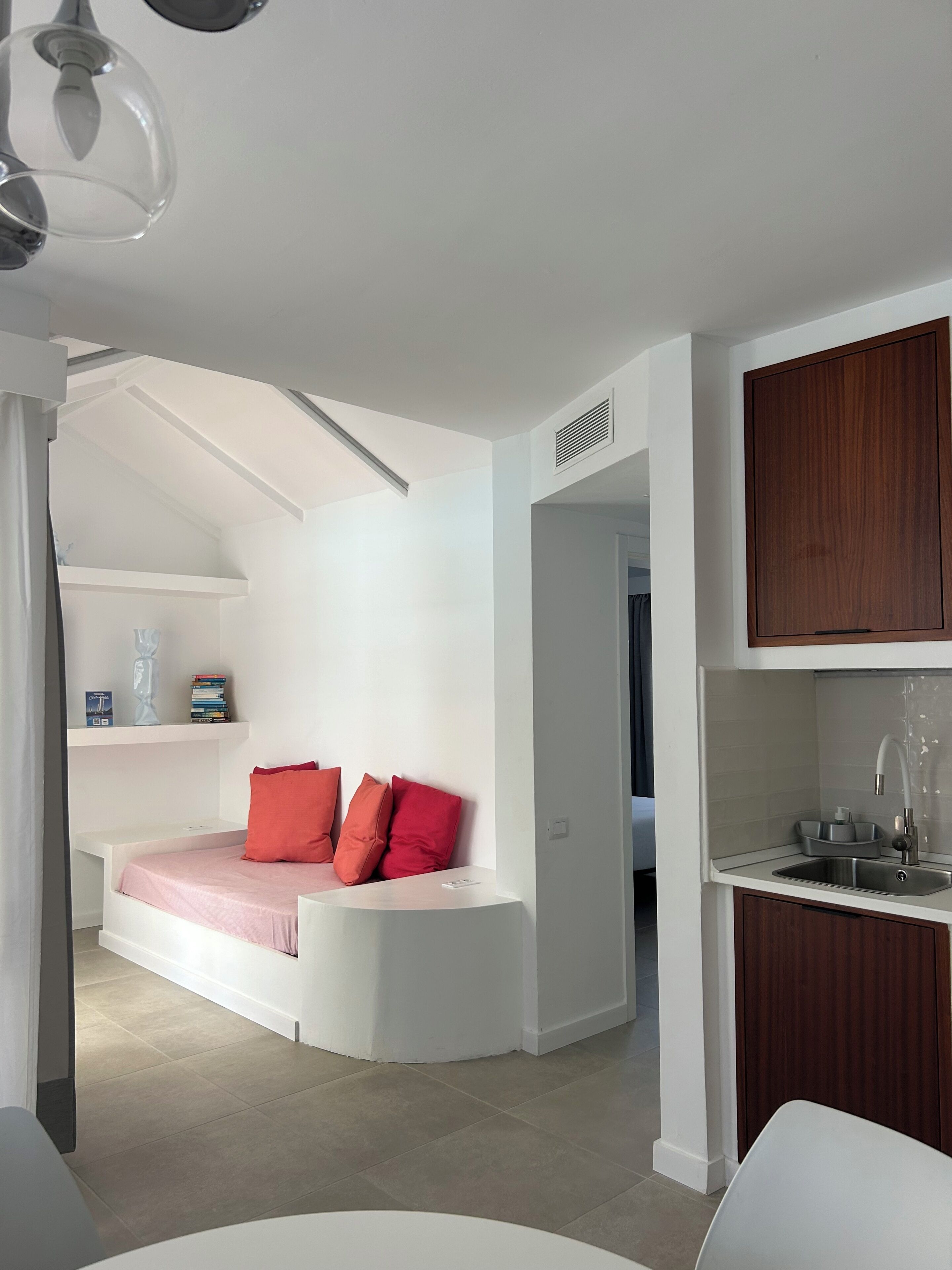 A modern interior with a white sofa, red cushions, a kitchen area with a sink, and a staircase leading to an upper level.