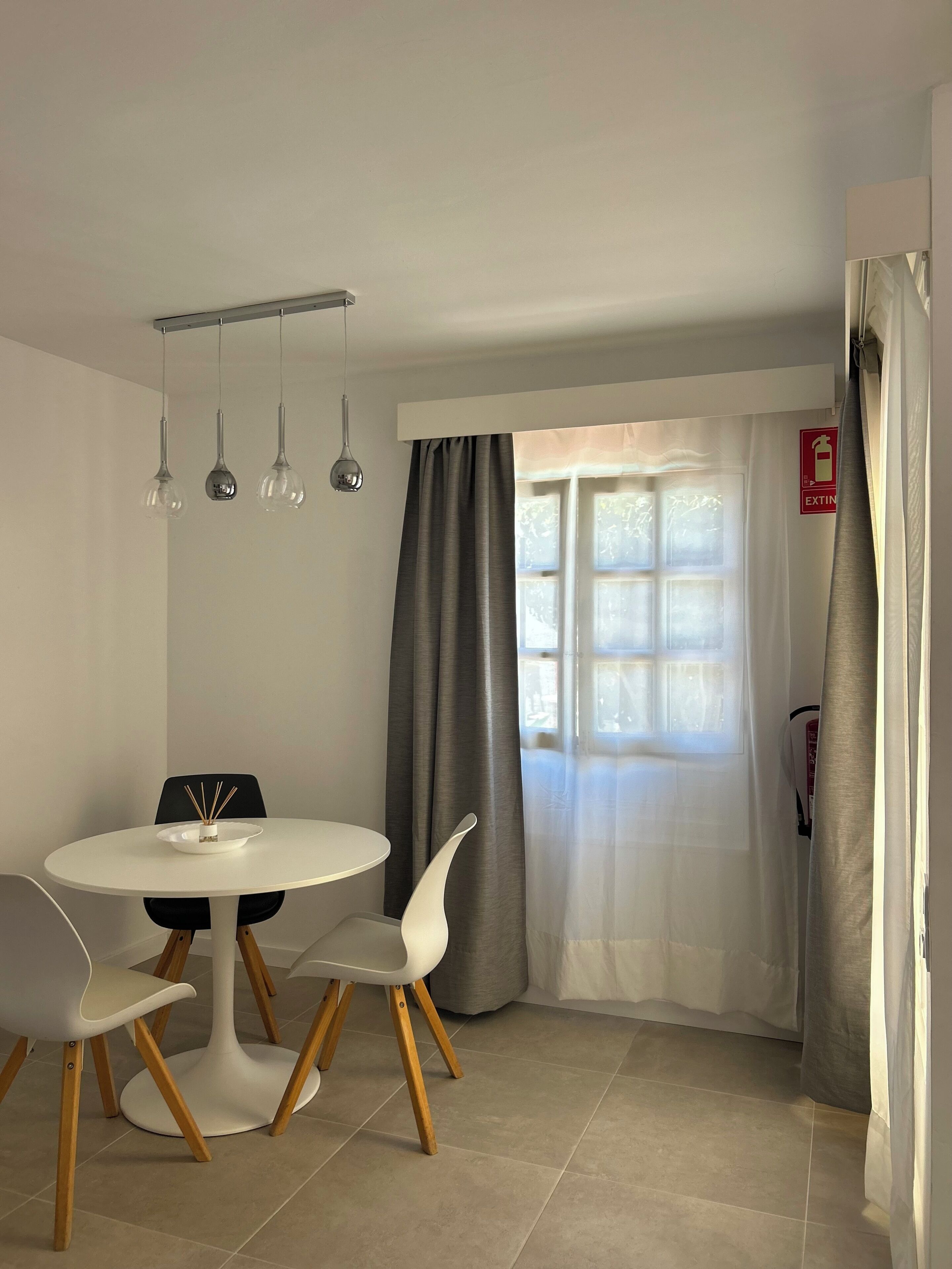 Un comedor moderno con una mesa redonda, sillas, luces colgantes, un ventanal con cortinas y una señal de salida.