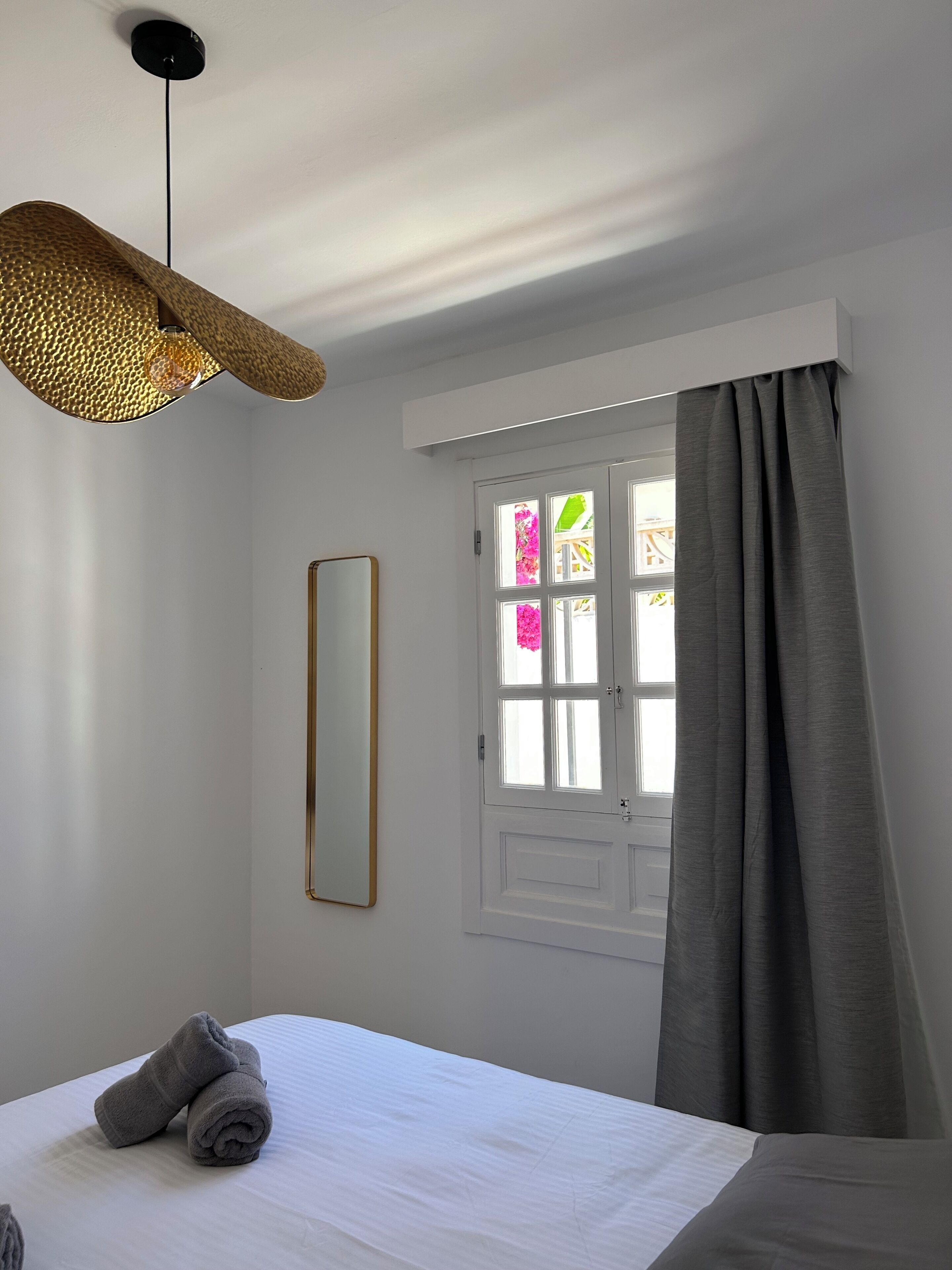Ein Schlafzimmer mit einem Bett, einer Hängelampe, einem Fenster mit Vorhängen und einem Spiegel an der Wand.