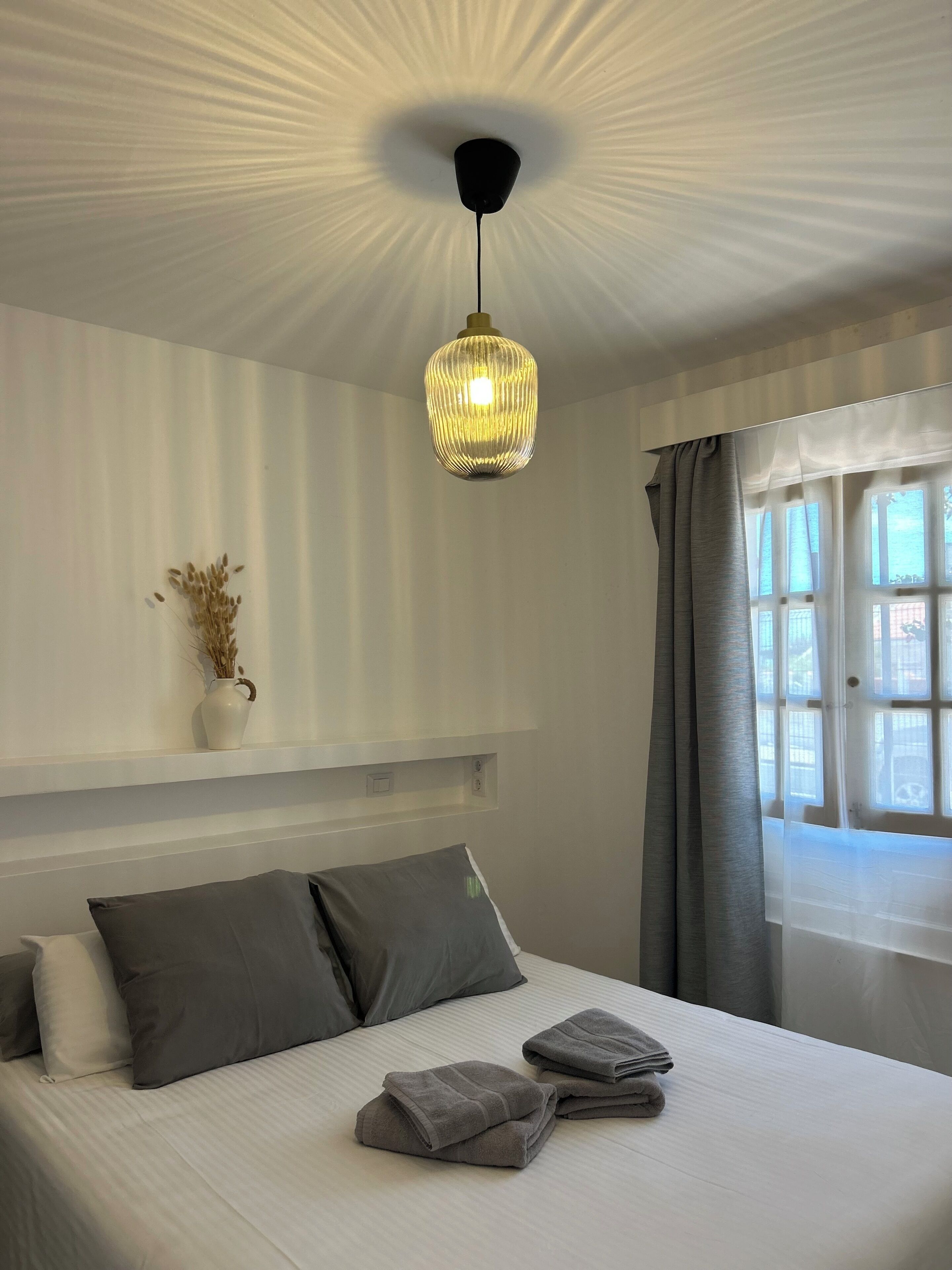 Une chambre à coucher avec un lit, des oreillers, une table de chevet avec un vase, un luminaire au plafond et une fenêtre avec des rideaux.