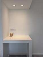 Un espacio pequeño y blanco, con techo bajo, una lámpara empotrada, un enchufe y un pequeño objeto decorativo sobre una superficie blanca.