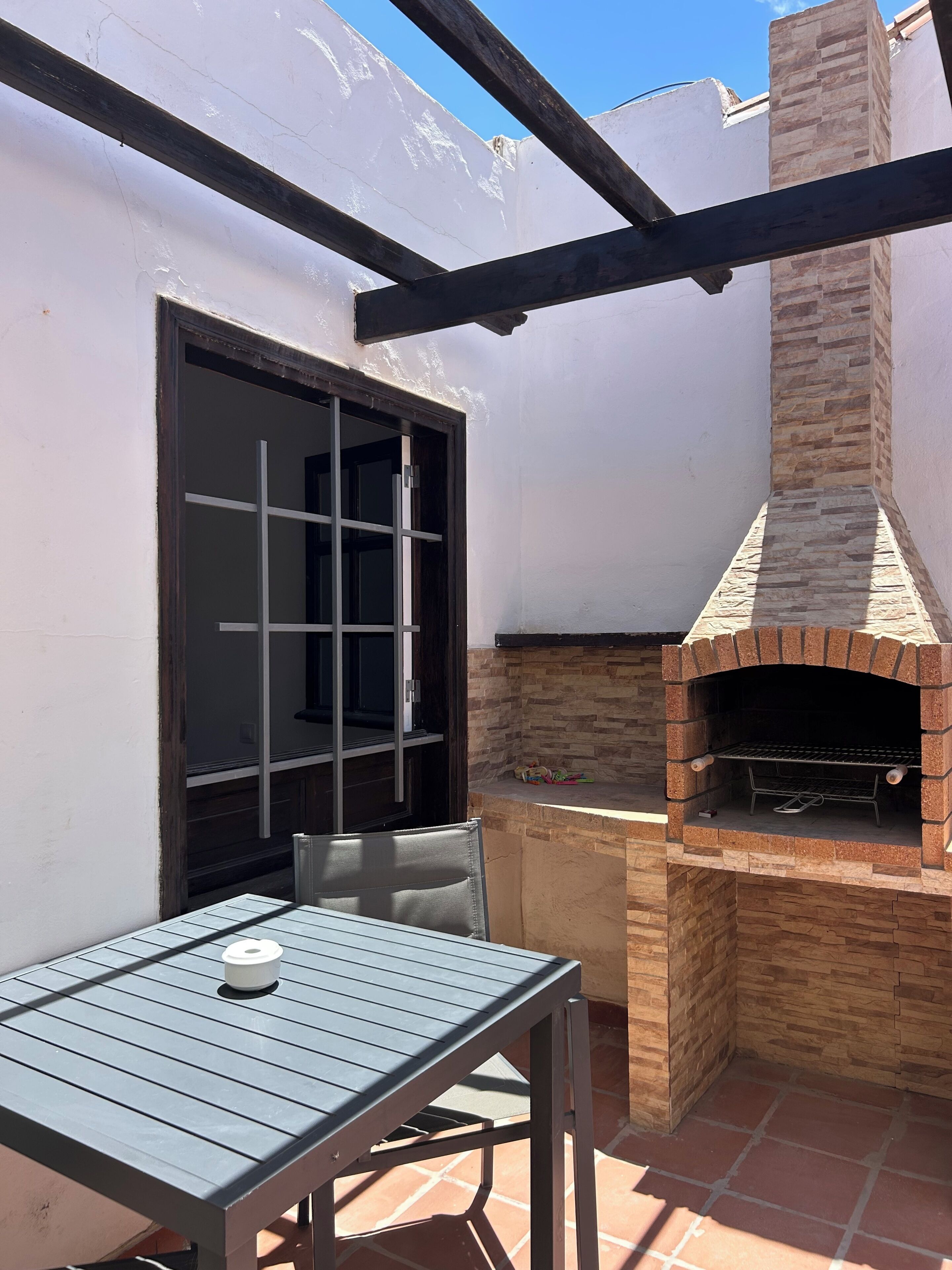 Een terras buiten met een tafel, stoelen, een bakoven en een raam met tralies.