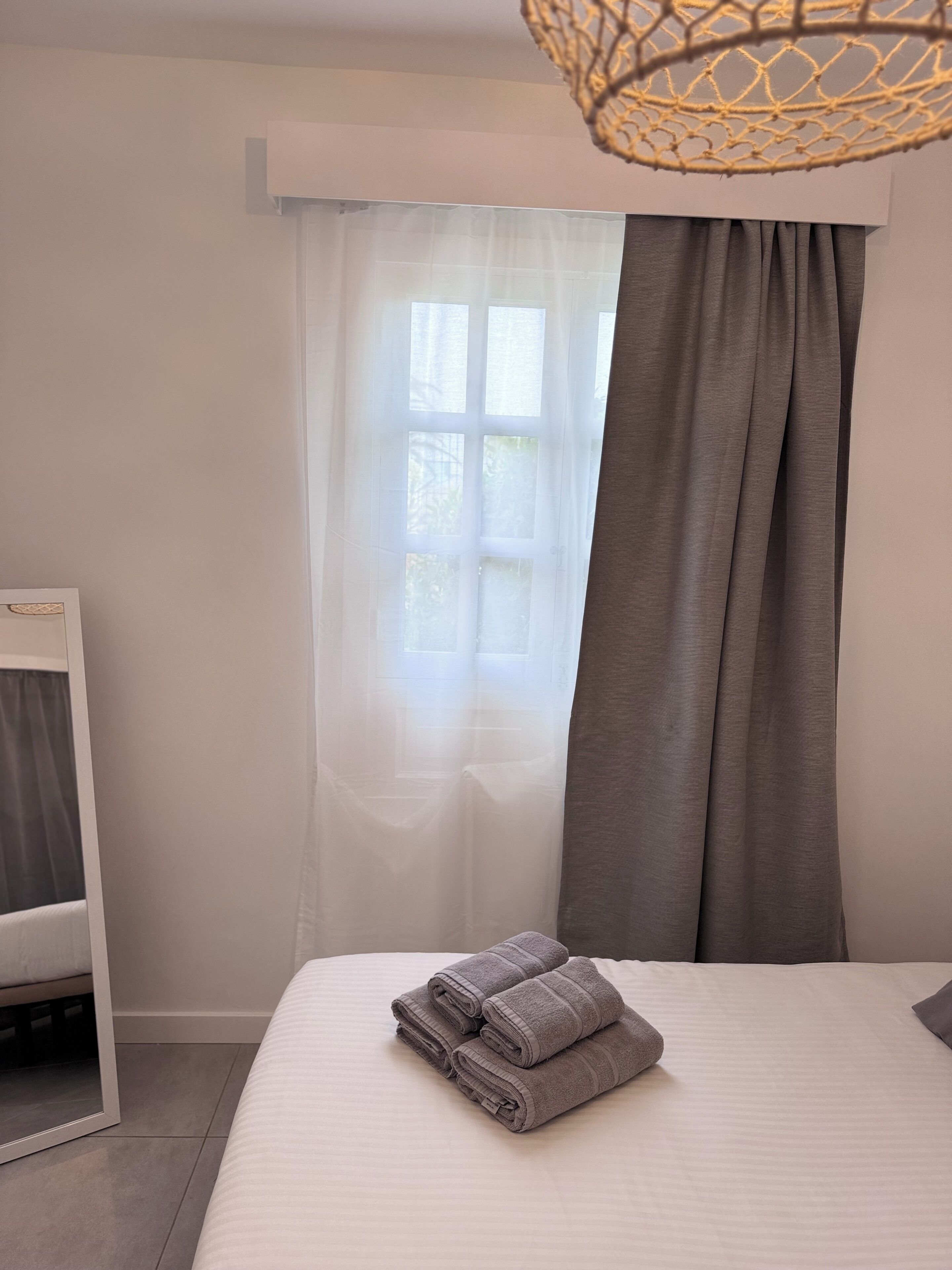 Una cama bien hecha con toallas dobladas, una mesita de noche, un espejo y una ventana con cortinas traslúcidas y un cortinaje más grueso.