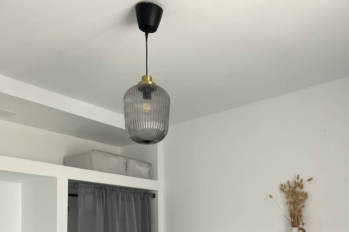 Ein Schlafzimmer mit einem Bett, grauen Vorhängen, einer Hängelampe und einer kleinen Vase mit Trockenblumen.