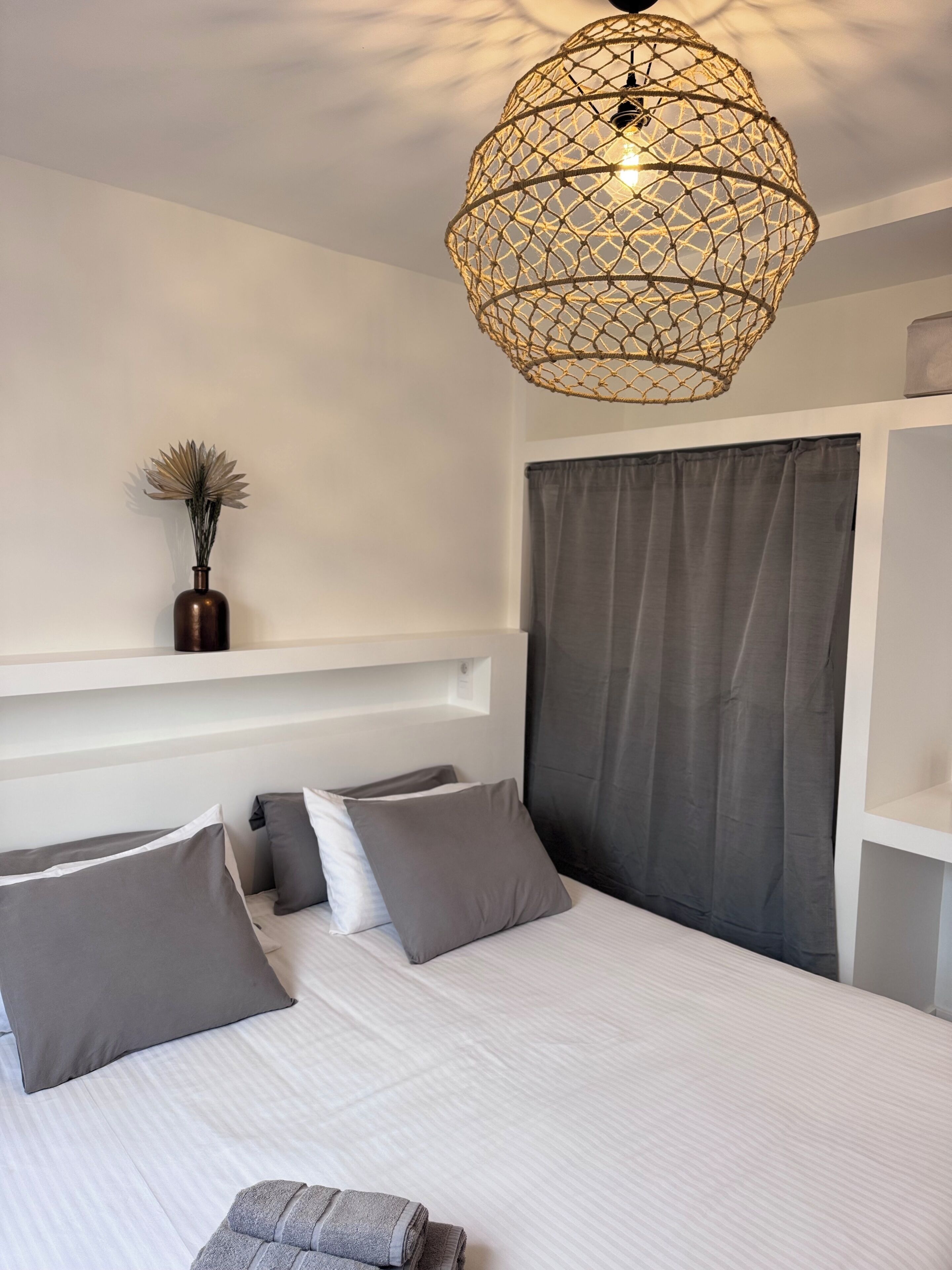 A bedroom with a bed, a hanging light fixture, a vase with dried flowers, and a window with curtains.