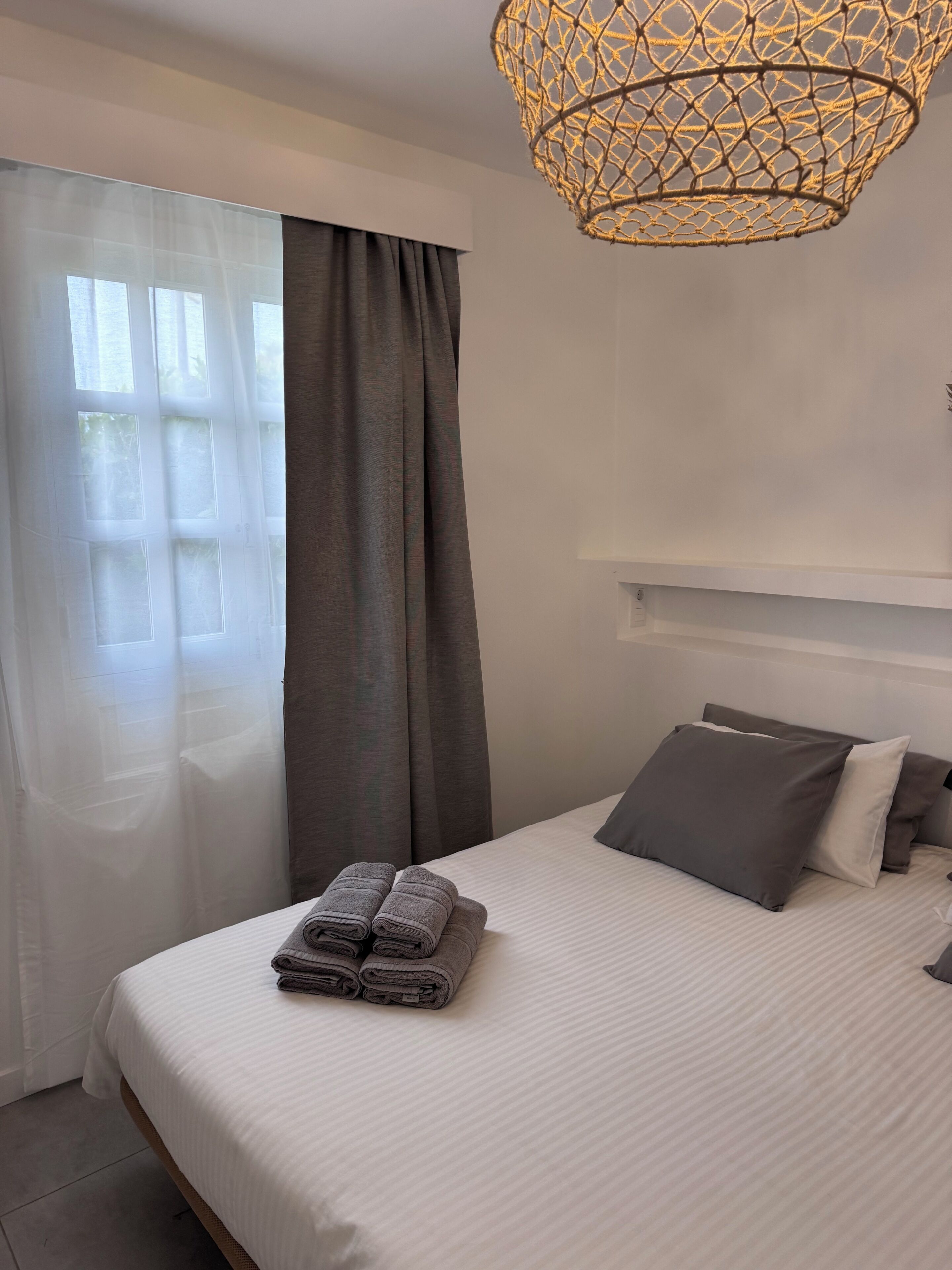 A neatly made bed with rolled towels, a hanging light fixture, and sheer curtains.