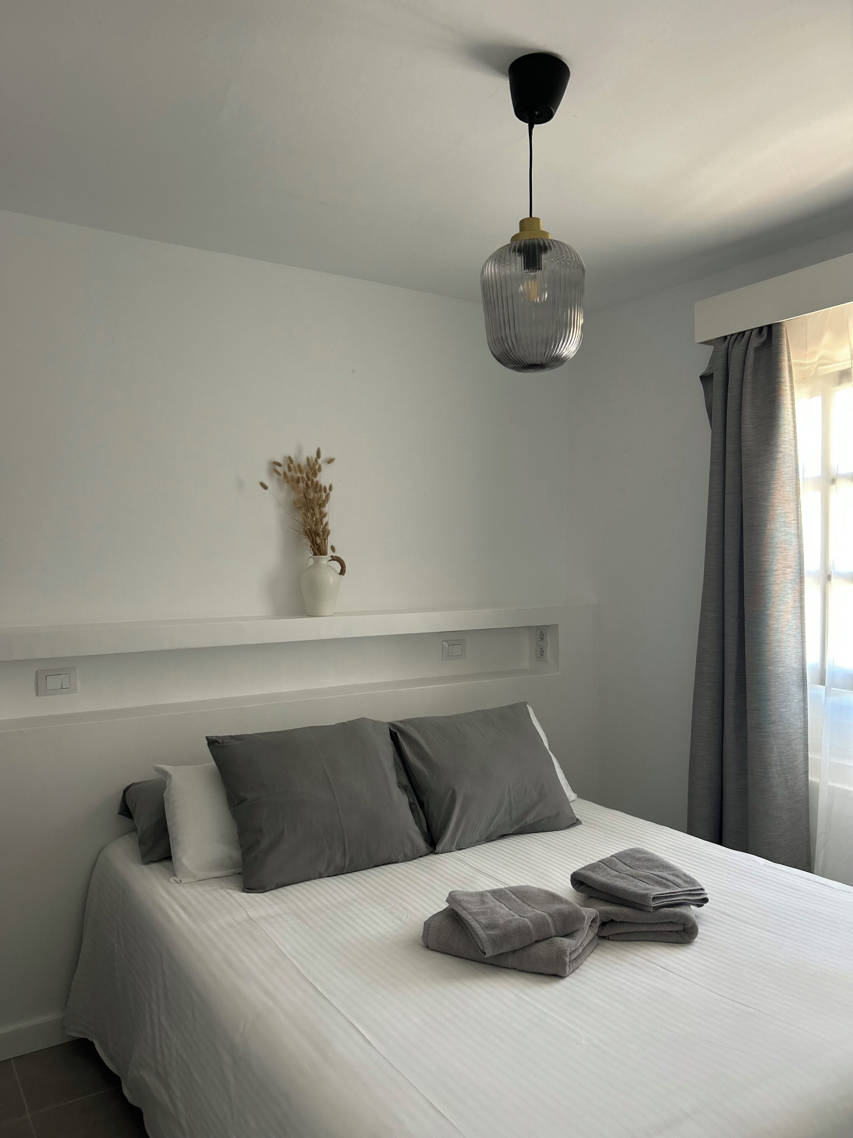 A bedroom with a bed, pillows, a bedside table, a vase with dried flowers, a hanging light fixture, and a window with curtains.