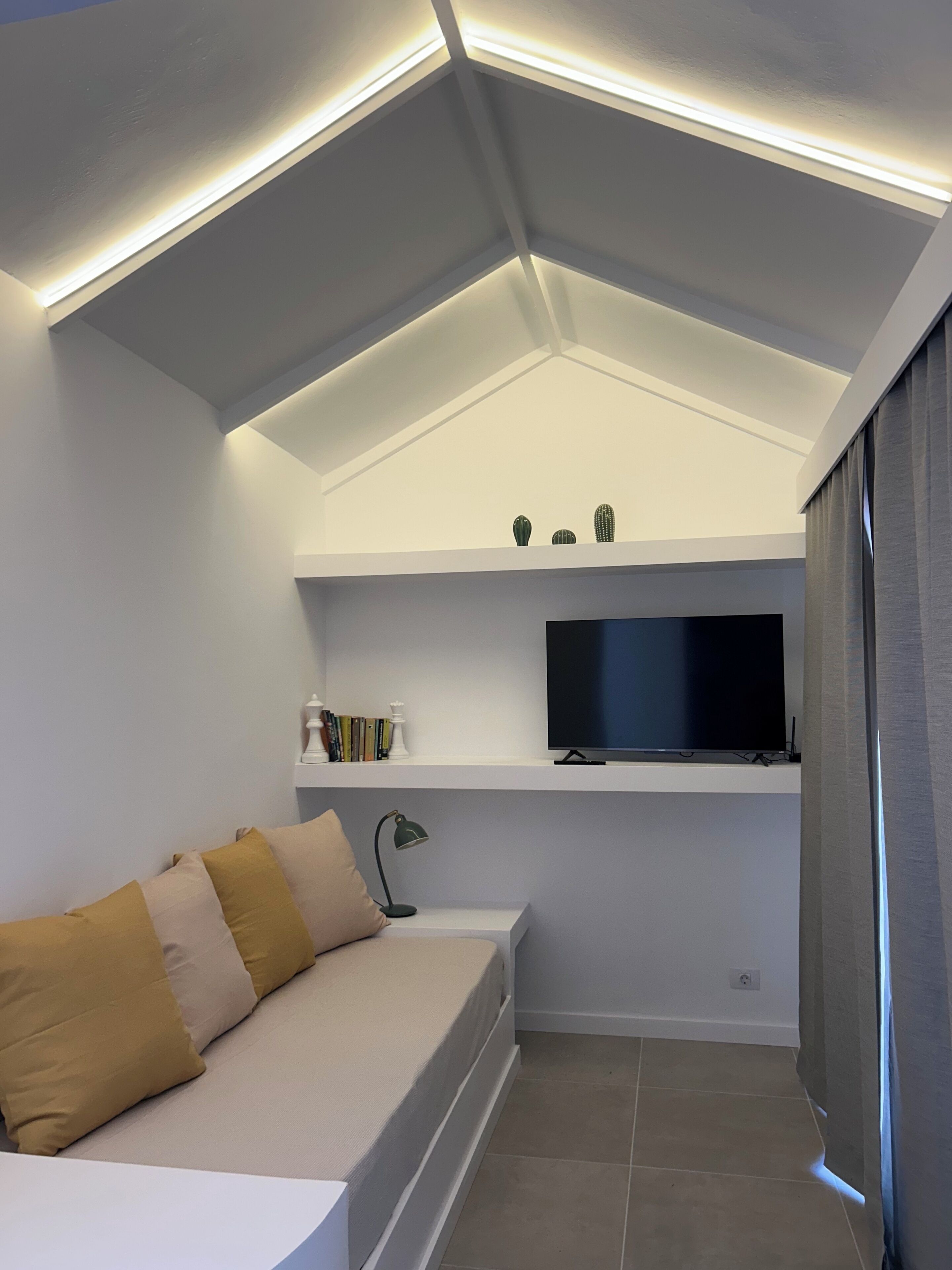 Quarto moderno com teto inclinado, TV de tela plana, sofá com almofadas e uma pequena estante com livros e objetos decorativos.