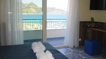 A hotel room with a bed, a desk, a television, and a view of the sea.