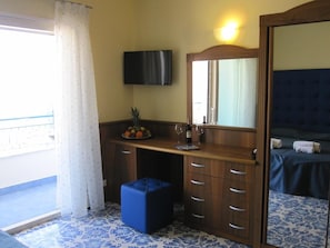 Habitació amb escriptori de fusta, mirall, televisió, llit amb capçalera blava i un otomà blau petit.
