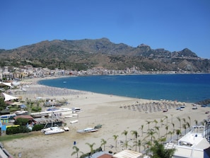 Un poble costaner amb platja, embarcacions i un paisatge de muntanyes al fons.