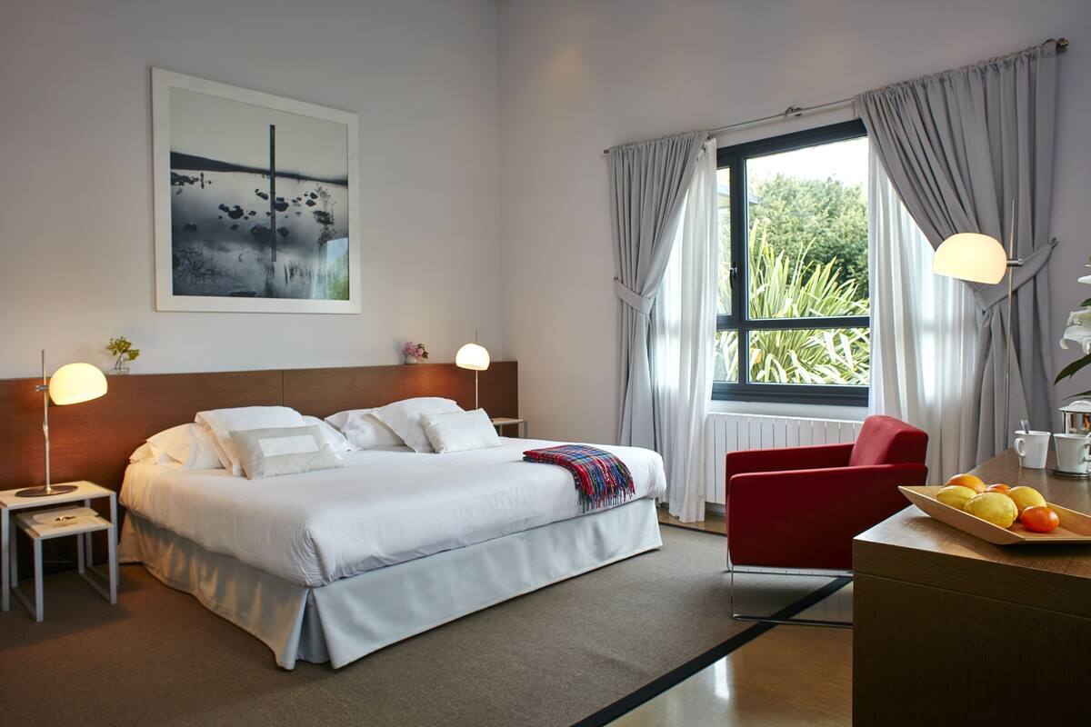 Ein Hotelzimmer mit einem Bett, einem roten Sessel, einem Esstisch mit Obst, einer Lampe, einem Gemälde und einem Fenster mit Vorhängen.