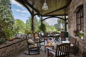 A covered patio with wooden furniture, stone walls, and a view of the garden.