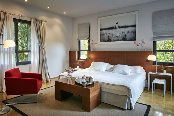 A modern hotel room with a large bed, a red armchair, a wooden coffee table, and a painting above the bed.