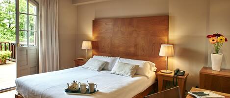 A neatly made bed with pillows, a bedside table with a lamp, a tray with a teapot and cups, and a vase with flowers.