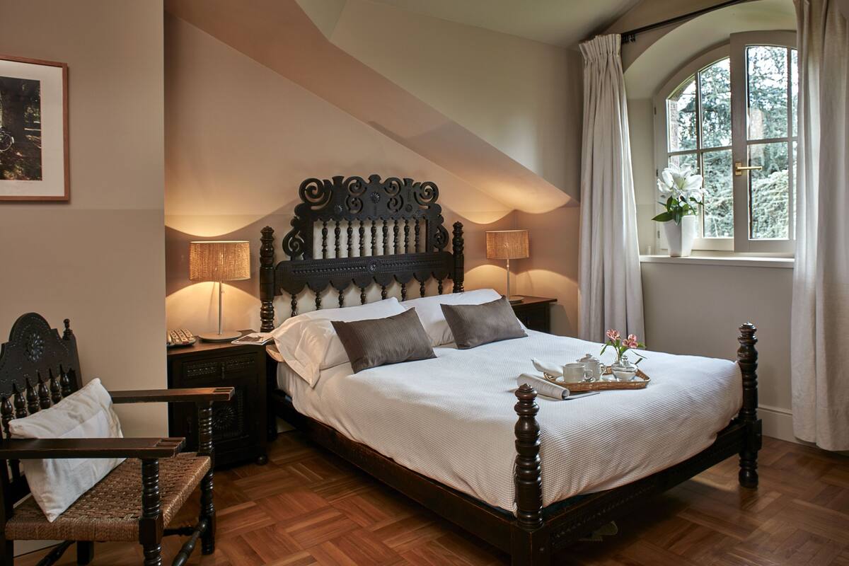Ein Schlafzimmer mit einem großen Bett, zwei Nachttischen, einem Sessel, einem Fenster mit Vorhängen und einem gerahmten Bild an der Wand.