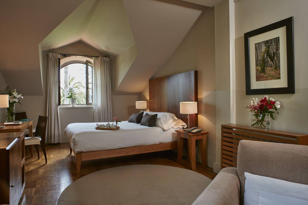Ein Hotelzimmer mit Bett, Nachttischen, Schreibtisch, Sessel, einer Couch, einem Fenster mit Vorhängen, einem gerahmten Bild an der Wand und einem Blumentopf.