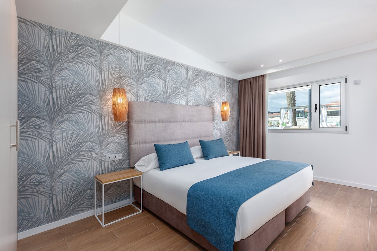 Une chambre moderne avec un grand lit, une table de chevet, deux lampes fixées au mur et une fenêtre donnant sur un beau paysage.