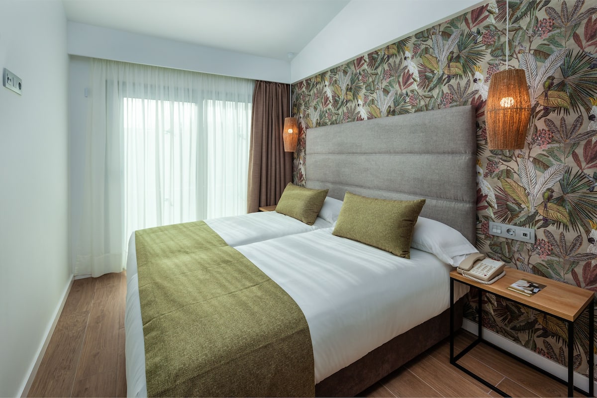Une chambre d’hôtel avec un grand lit, une table de chevet avec un téléphone et un mur recouvert d’un papier peint aux motifs tropicaux.