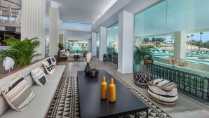 A spacious living area with a large glass wall offering a view of a pool and outdoor area. There is a black table with decorative bottles and a patterned rug. - Kumara Serenoa by Lopesan Hotels (San Bartolomé de Tirajana)