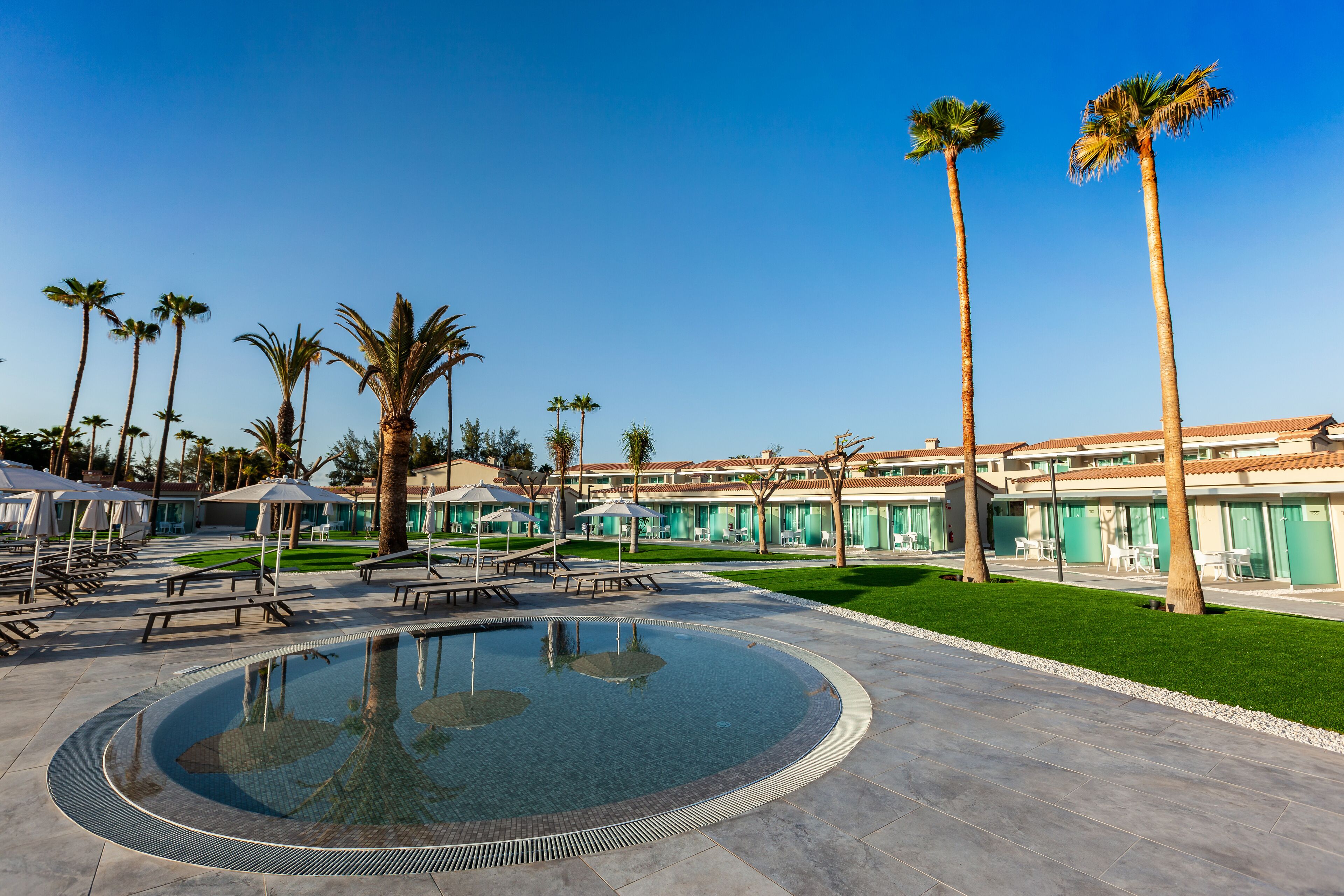 A pool area with lounge chairs and umbrellas, surrounded by palm trees and a building with a tiled roof.