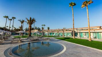 A pool area with lounge chairs and umbrellas, surrounded by palm trees and a building with a tiled roof.