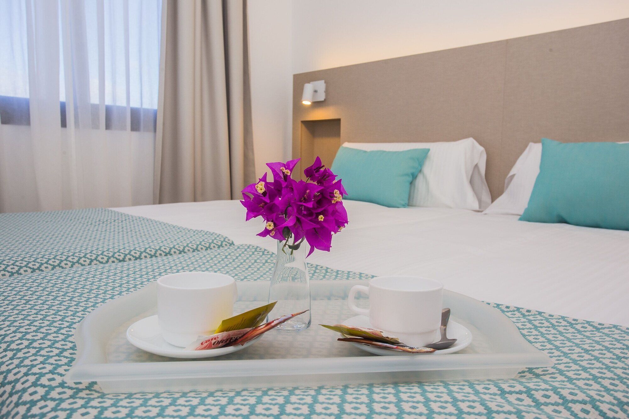 Ein Hotelzimmer mit einem Bett, einem Tablett mit zwei Tassen und einer Blumenvase, sowie einem Fenster mit Vorhängen.
