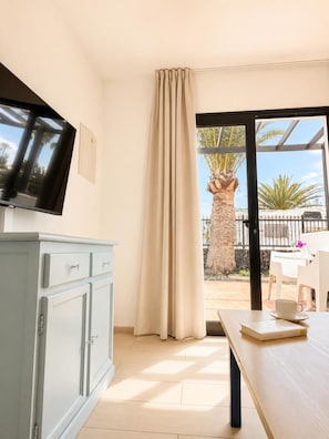 A room with a television, a dining table with a cup, and a view of palm trees through a window. - Los Tulipanes (Tias)