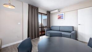 Un salon moderne avec une table ronde, deux chaises, un canapé gris, un luminaire suspendu et une porte-fenêtre coulissante donnant sur un balcon.
